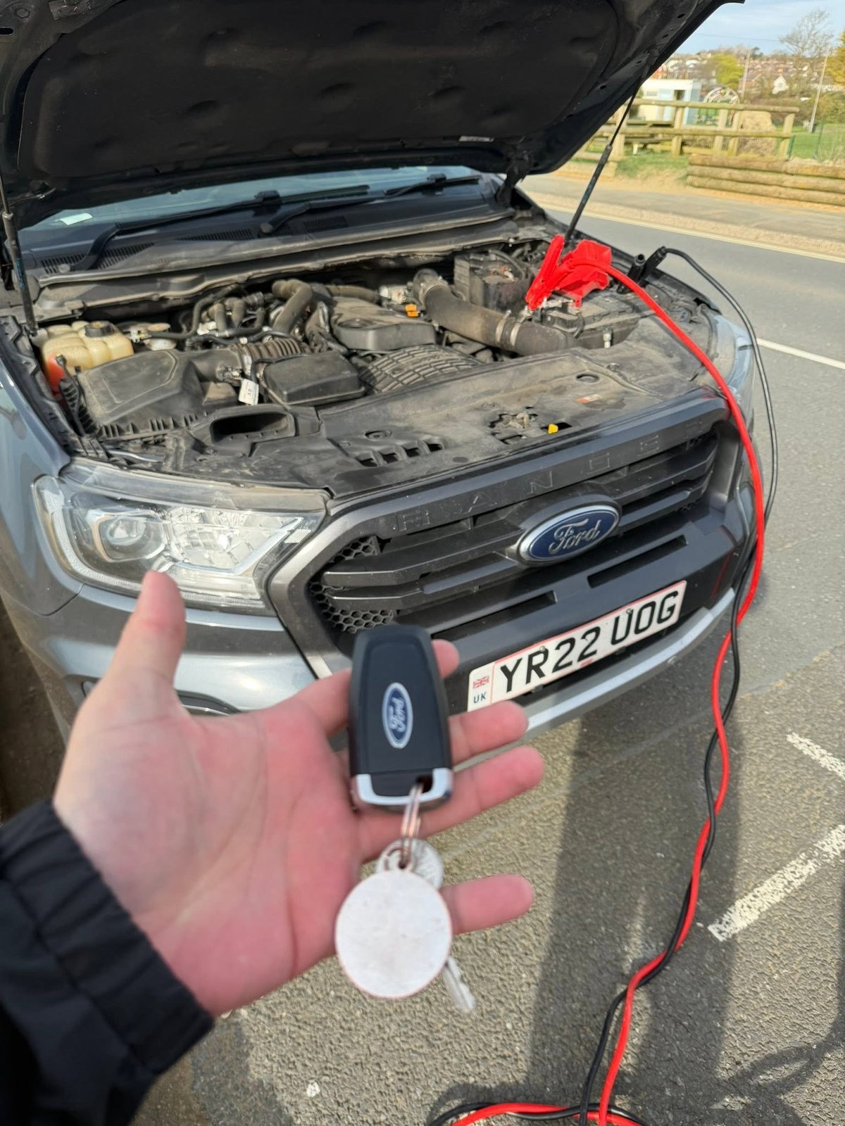 Hand holding Ford car key with red charging cables connected to open vehicle engine bay
